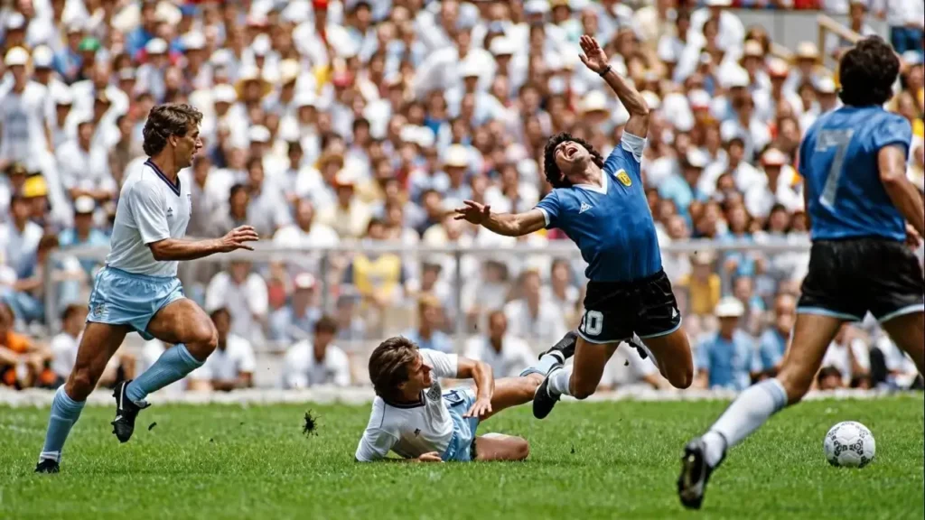 Diego Armando Maradona cae tras una infracción del defensor inglés Terry Fenwick, mientras Kenny Sansom se acerca a la jugada durante los cuartos de final del Mundial de México 1986, disputados el 22 de junio de 1986 en el Estadio Azteca de la Ciudad de México. La imagen corresponde al histórico triunfo de Argentina por 2-1 sobre Inglaterra, partido en el que Maradona marcó "La Mano de Dios" y "El Gol del Siglo".