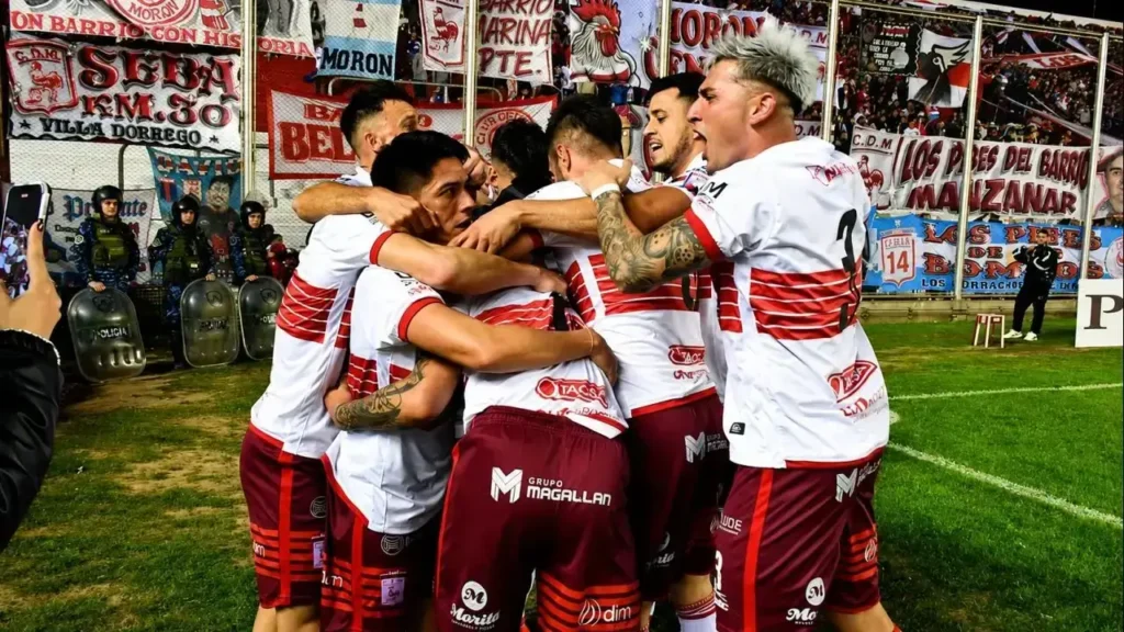 Los jugadores de Deportivo Morón celebran abrazados sobre el campo de juego tras una acción decisiva, en una imagen que refleja la unión del plantel y el fervor de la hinchada. Al fondo se destacan las banderas de la Peña Villa Dorrego, Barrio Marina, Los Pibes del Barrio Manzanar y otras agrupaciones que acompañaron al Gallo desde la tribuna.