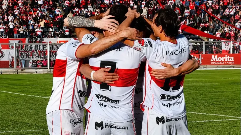 Los jugadores de Deportivo Morón celebran abrazados frente a una tribuna repleta en el Nuevo Francisco Urbano. La imagen refleja la unión del plantel y el respaldo de la hinchada, mientras luce la camiseta titular 2026, con Milo J como patrocinador principal en el frente de la casaca.