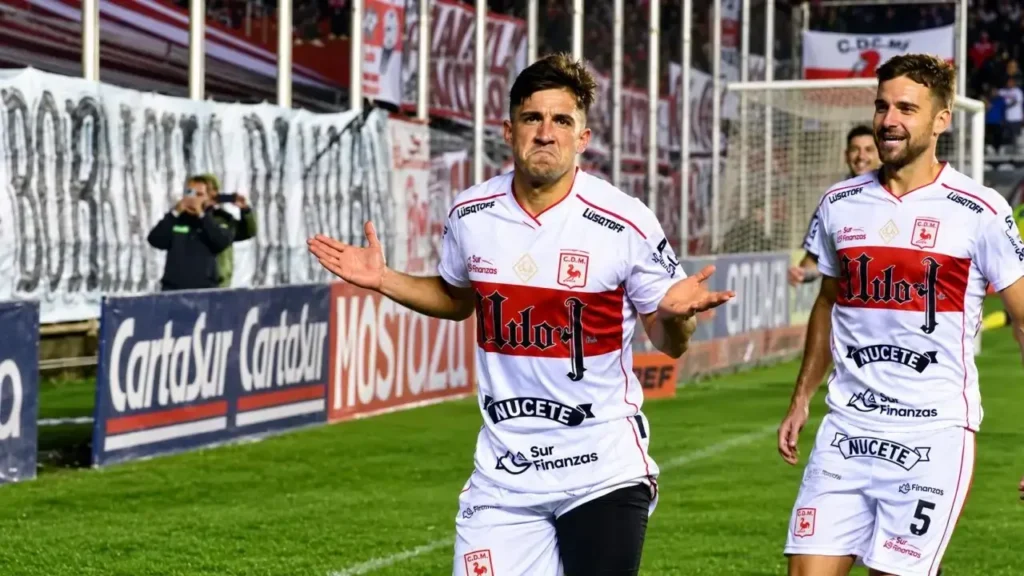 Dos futbolistas de Deportivo Morón celebran una conquista en el Nuevo Francisco Urbano con la camiseta titular que lleva a Milo J como patrocinador principal. El festejo refleja el clima de comunión entre el plantel y la hinchada en una noche de Primera Nacional.