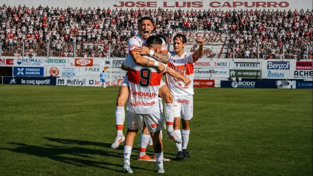Festejo a pura emoción. Los jugadores de Deportivo Morón celebran uno de los goles del triunfo en el Nuevo Francisco Urbano, con un abrazo que resume la entrega del equipo y el respaldo de un estadio colmado. Al fondo, la popular del Gallo acompaña la celebración con un imponente marco de banderas e hinchas.