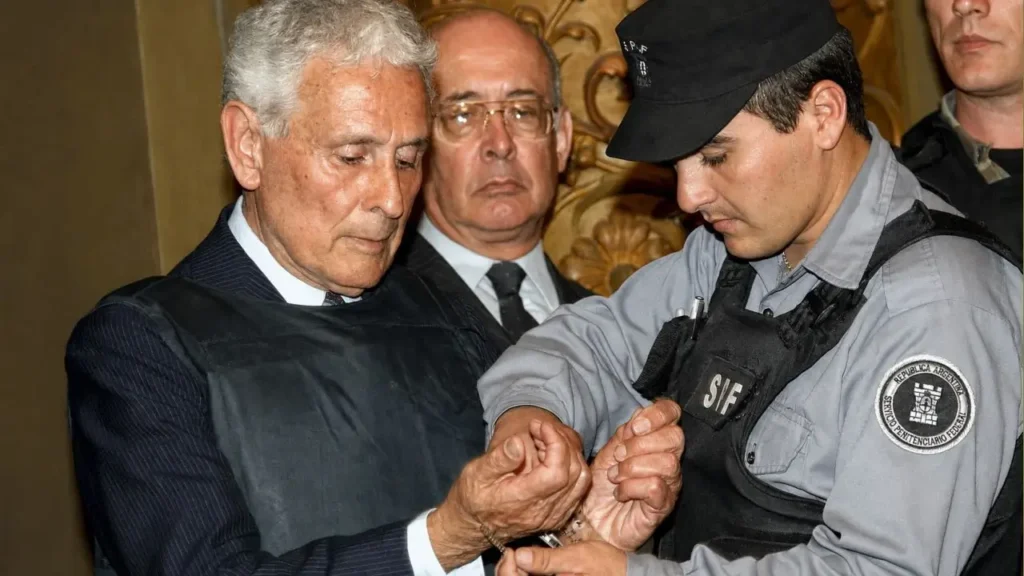 Miguel Osvaldo Etchecolatz es esposado por un agente del Servicio Penitenciario Federal en el Tribunal Oral Federal N.º 1 de La Plata, el 19 de septiembre de 2006, después de ser condenado a reclusión perpetua por homicidios, secuestros y torturas cometidos durante la última dictadura, delitos que el tribunal consideró de lesa humanidad perpetrados en el marco de un genocidio.