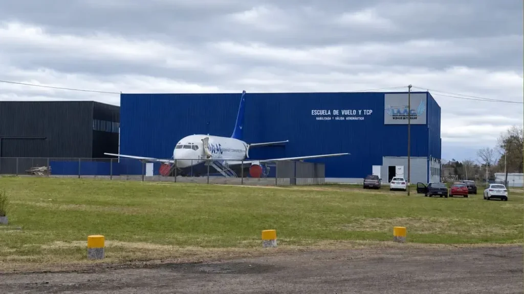 Fachada principal de la Escuela de Vuelo y TCP de Wings Aerospace Academy. El gran hangar azul sirve de fondo para un avión comercial de entrenamiento, destacando la infraestructura dedicada a la formación de pilotos y tripulantes de cabina de pasajeros (TCP), bajo la leyenda "Habilitada y Válida Aeronáutica".