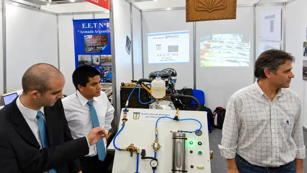Durante la exposición, un estudiante señala partes del generador de hidrógeno para motores térmicos mientras otro acompaña la presentación. La instancia de intercambio también forma parte del aprendizaje: comunicar el proceso y responder consultas es parte del trabajo técnico que los alumnos llevan de la escuela a la feria.