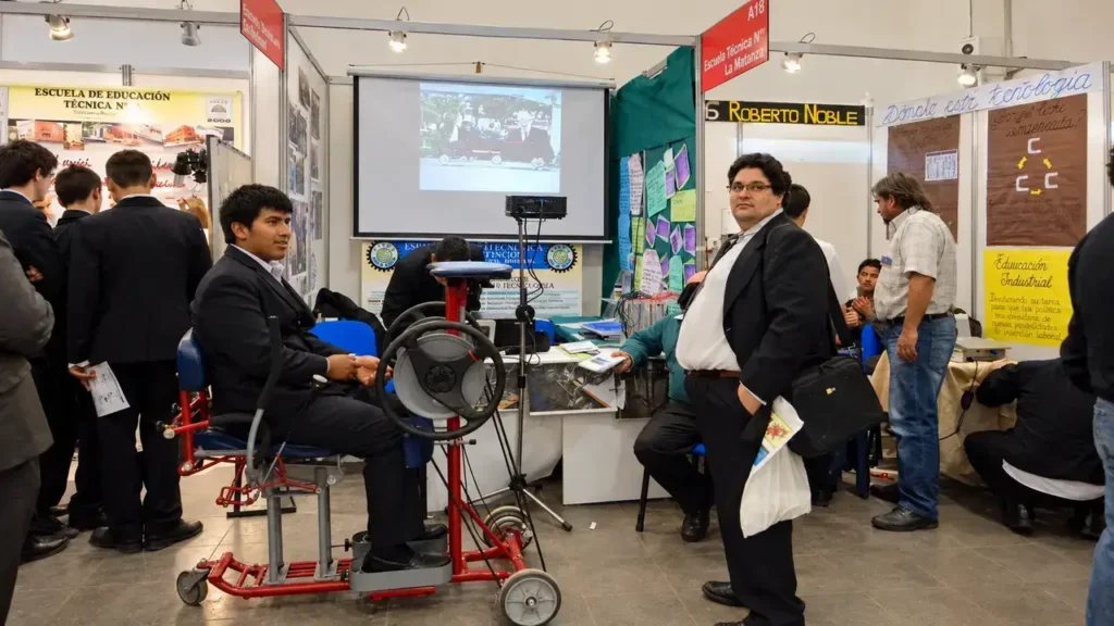 Un alumno prueba un simulador de conducción en el espacio de una escuela técnica, mientras visitantes y docentes recorren el stand. La propuesta suma una dimensión interactiva a la exposición y acerca al público a los proyectos que se desarrollan en las aulas y talleres.