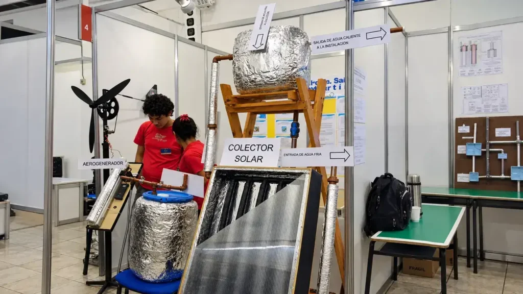 Estudiantes presentan un colector solar y un aerogenerador, junto con las conexiones que permiten mostrar el recorrido del agua en el sistema térmico. Los carteles distinguen la entrada de agua fría y la salida de agua caliente, y ayudan a explicar cómo se organiza el prototipo.