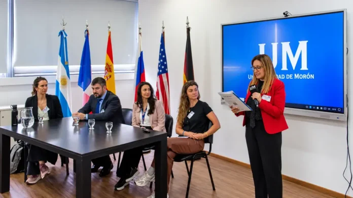 La Universidad de Morón realizó una nueva edición de la Expo Internacional UM. La actividad se desarrolló en el Hall del Bicentenario, en la sede de Machado 854, y formó parte de la estrategia de internacionalización de la institución. En la foto, una expositora presenta ante un panel en la Universidad de Morón. La actividad se desarrolla en una sala con banderas de distintos países y una pantalla institucional de la UM, en un encuentro dedicado al intercambio académico y cultural.