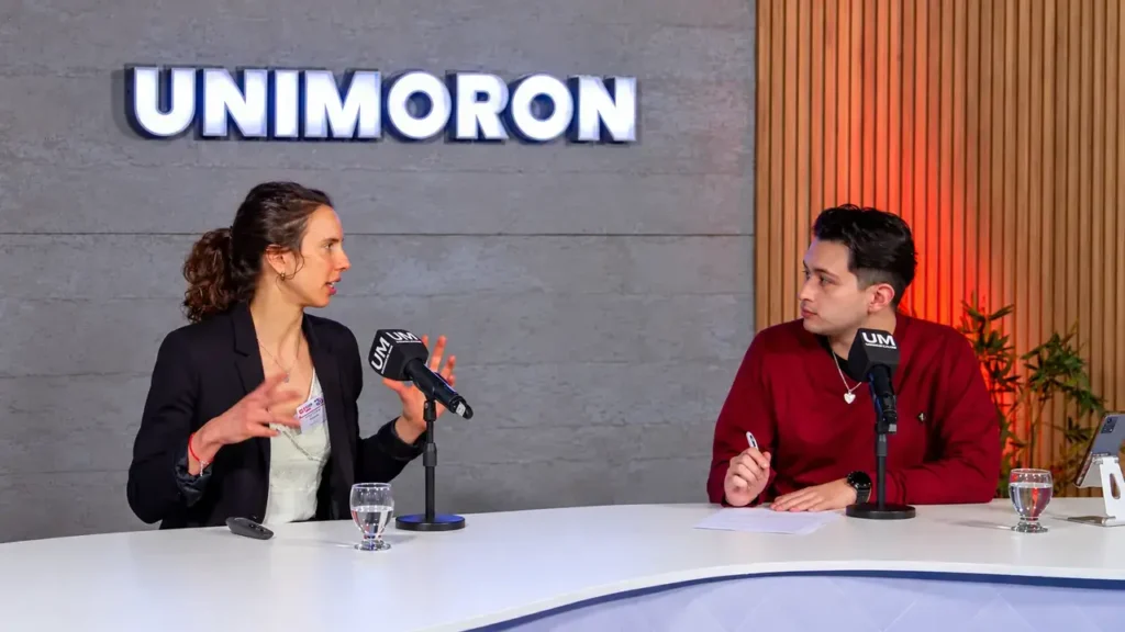 Una entrevistada comparte sus ideas durante una conversación en vivo. Frente a ella, un joven toma notas mientras ambos participan de una transmisión o entrevista con micrófonos identificados con la Universidad de Morón.