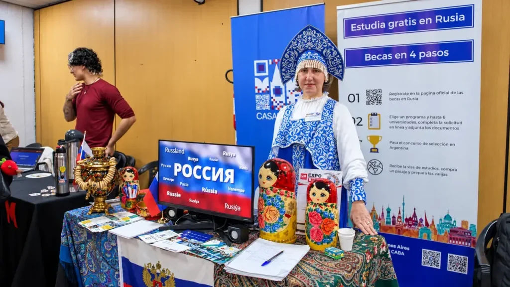 El stand de Rusia presenta su cultura y oportunidades de estudio. Una expositora muestra objetos tradicionales, publicaciones y material sobre becas universitarias, mientras un cartel explica los pasos para postularse a programas de formación en ese país.