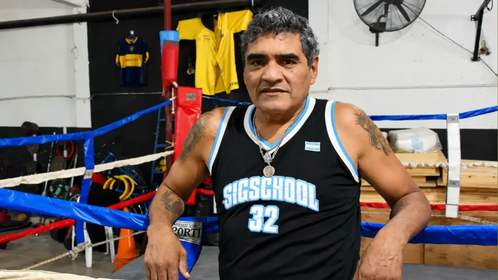 Jorge “Locomotora” Castro posa junto al ring de un gimnasio, con una camiseta deportiva y los guantes apoyados sobre las cuerdas. El excampeón mundial construyó una extensa trayectoria de 144 combates profesionales, con 130 victorias, 90 de ellas por nocaut; murió a los 59 años tras descompensarse en el Bingo de Morón.